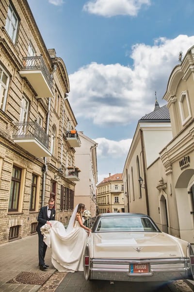 Vestuvių fotografas Vilniuje: jaunikis padeda nuotakai su ilga uodega įlipti į kreminį Cadillac senamiesčio gatvėje
