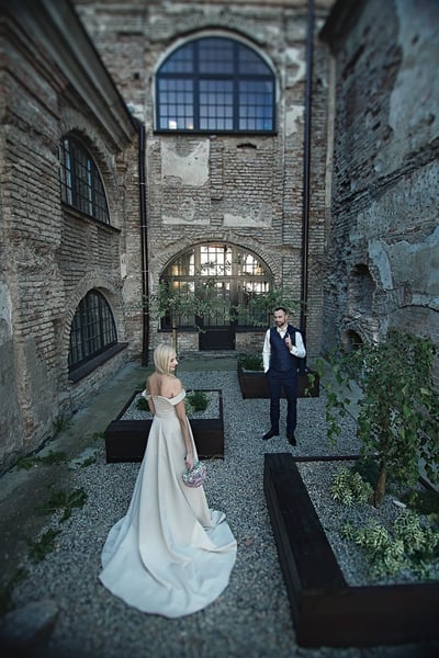 Vestuvių fotosesija: nuotaka su šydu tamsiame senos architektūros kieme, jaunikis fone