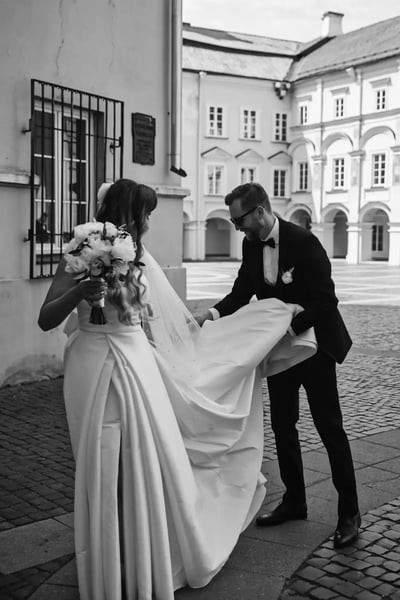 Vestuvių fotografas Vilniuje: jaunikis padeda nuotakai sutvarkyti suknelės uodegą senamiesčio kieme, nespalvota nuotrauka