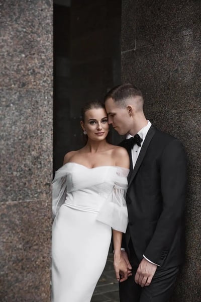 Vestuvių fotografas Kaune: nuotaka baltoje suknelėje su skaidriomis rankovėmis ir jaunikis juodu kostiumu prie granito sienos